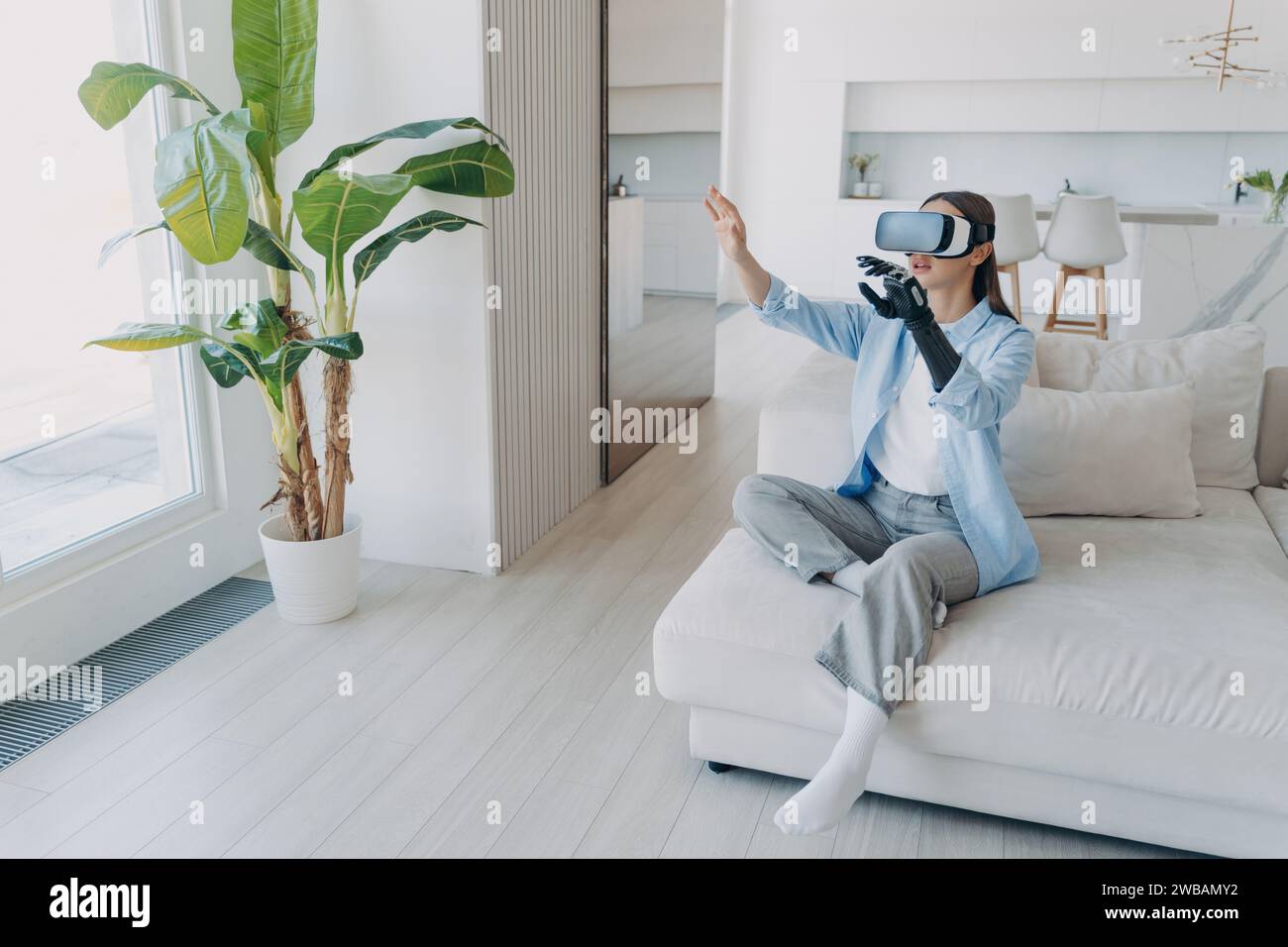 Amazed woman with prosthetic arm experiencing virtual reality, gesturing while seated on a couch in a sunlit room Stock Photo