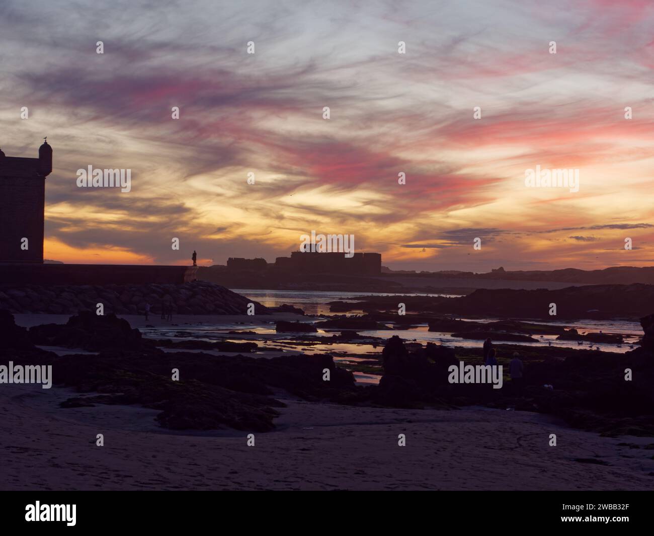 Dramatic sunset over the rocky coastline and fort area as someone in silhouette stands on the city walls, in Essaouira, Morocco. 9th Jan 2024 Stock Photo