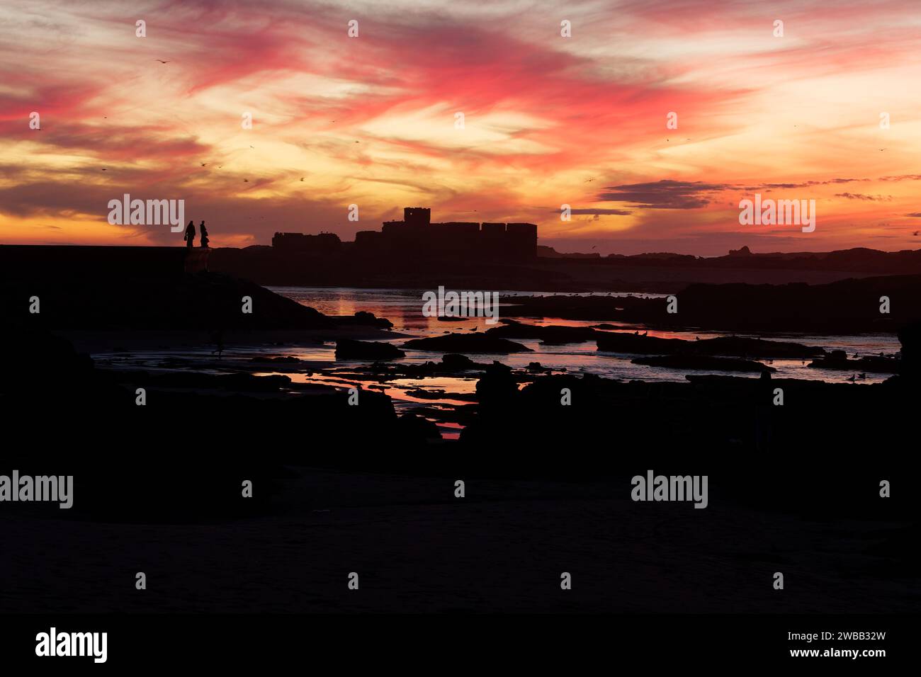 Dramatic sunset over the rocky coastline and fort area as people in silhouette stands on the city walls, in Essaouira, Morocco. 9th Jan 2024 Stock Photo
