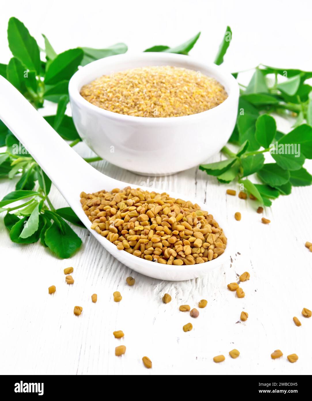 Fenugreek seeds in a spoon and ground spice in a bowl with leaves on background of light wooden board Stock Photo