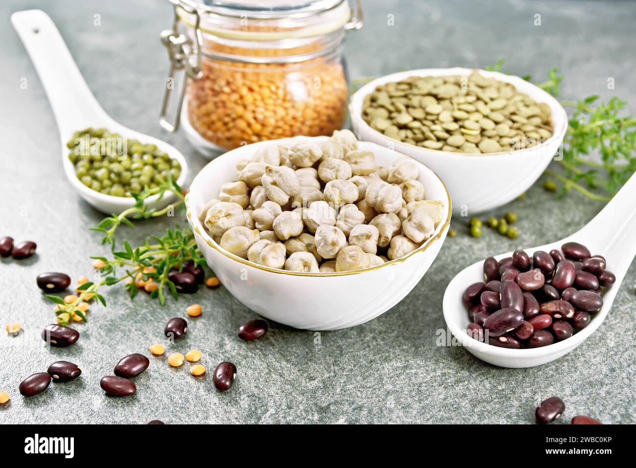 Green lentils and chickpeas in bowls, ruby haricot and mung beans in spoons, red lentils in a glass jar, thyme sprigs on the background of a stone tab Stock Photo