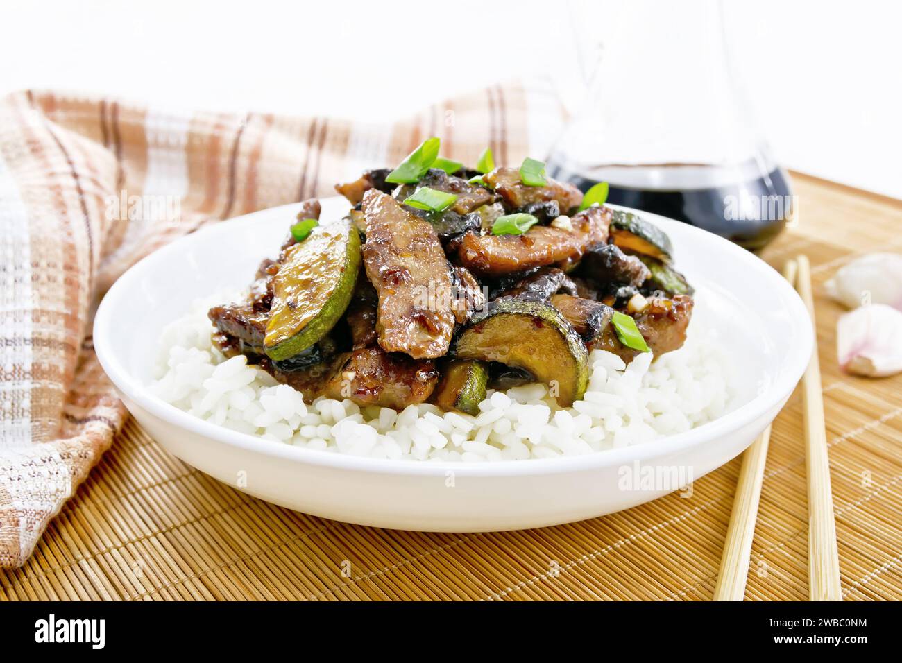 Chinese stir-fried dish of chicken meat with zucchini, mushrooms, soy sauce and rice garnish in a plate on a bamboo napkin, a towel and garlic on wood Stock Photo