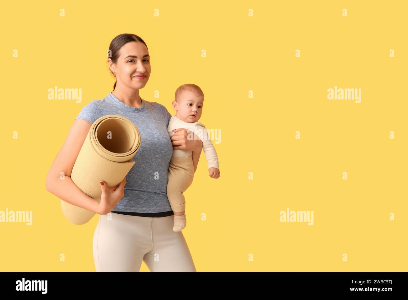 Beautiful young sporty woman with yoga mat holding her little baby on
