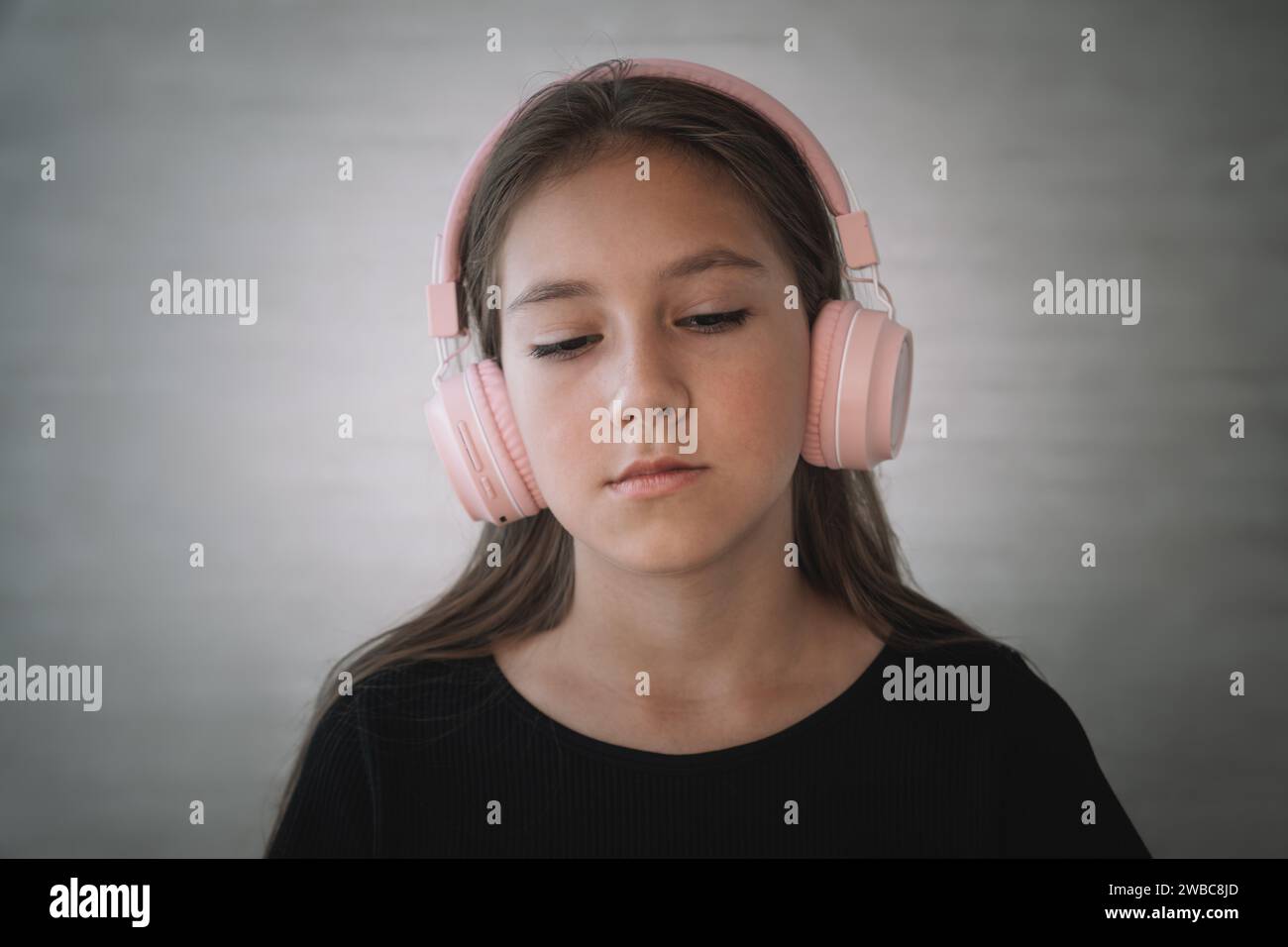 Serious displeased Caucasian kid girl wearing black dress being angry wears stereo headphones listens music Stock Photo
