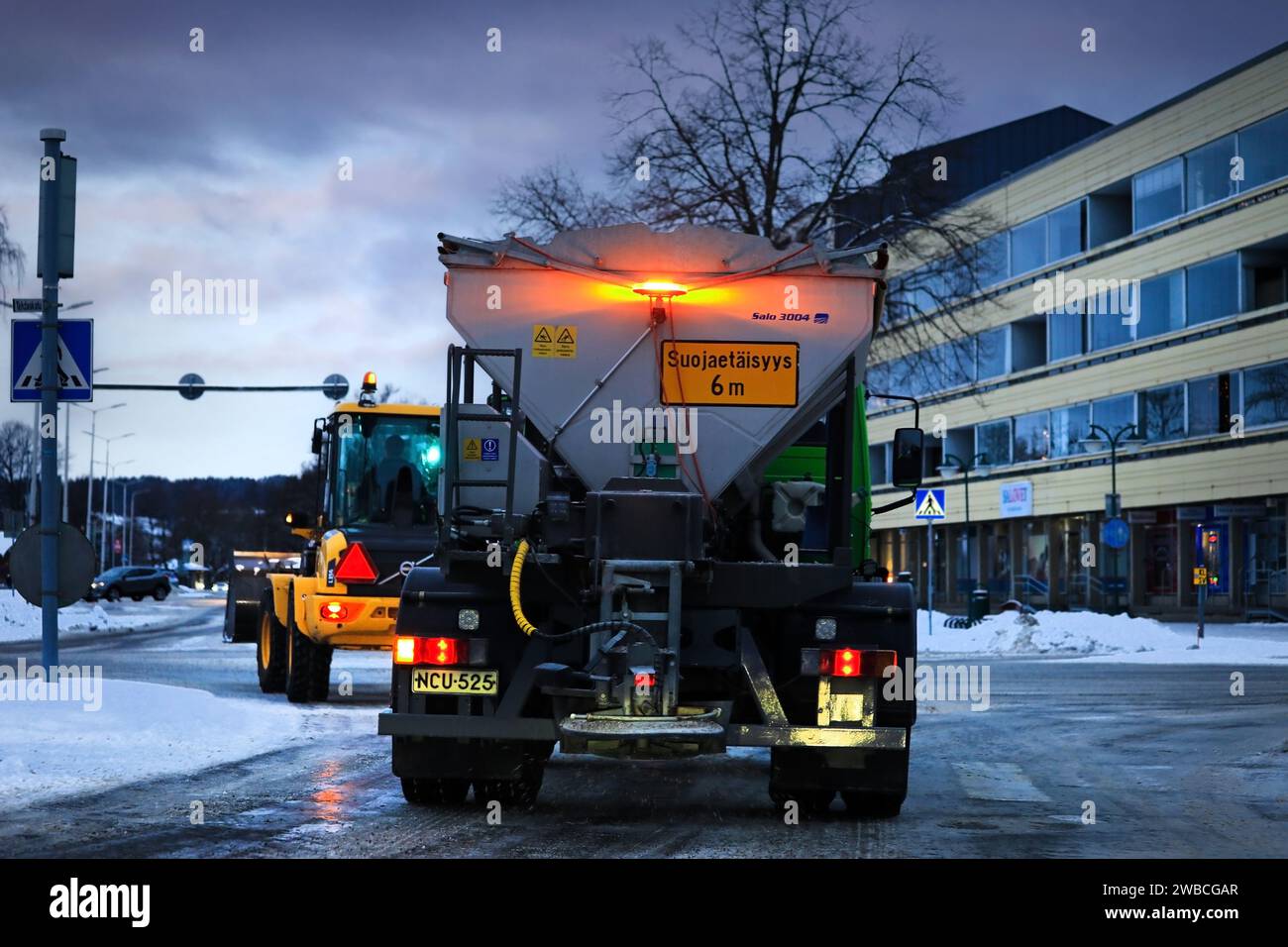 Gritting truck spreading grit on street with snow tractor ahead turning left at traffic lights. Rear view. Salo, Finland. December 29, 2023. Stock Photo