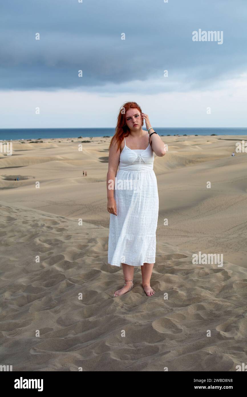 A young girl stands in a white dress in the sand dunes under a cloudy sky Stock Photo
