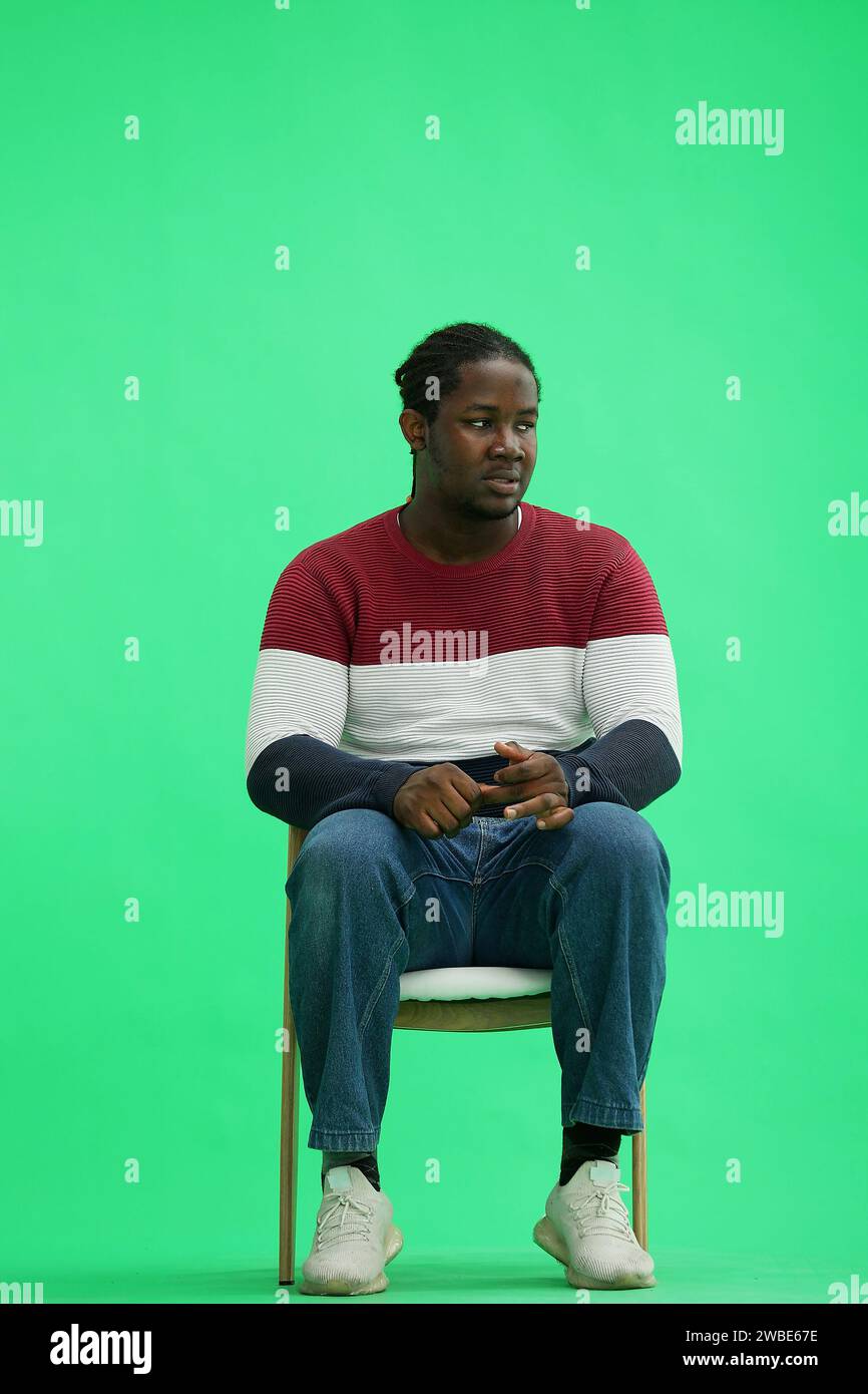 A man in a multi-colored jacket with a backpack on a green background sits on a chair Stock Photo