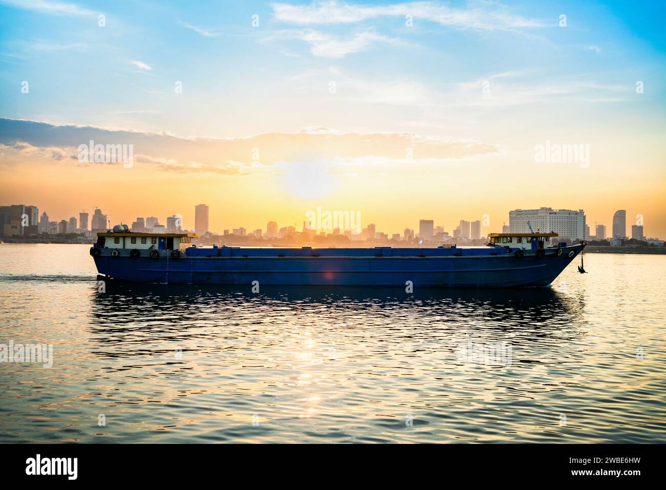 Cargo ship and dry bulk vessel at sunset. Export or import in Asia. Grain or wheat transportation. Phnom Penh city skyline in the background. Stock Photo