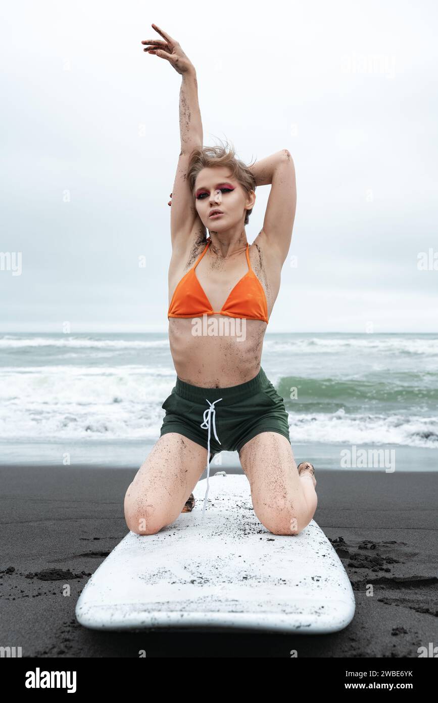 Flexibility woman surfer is posing on her knees on surfboard on sandy beach. female surfer looks amazing as sports fashion model, raising one hand up Stock Photo