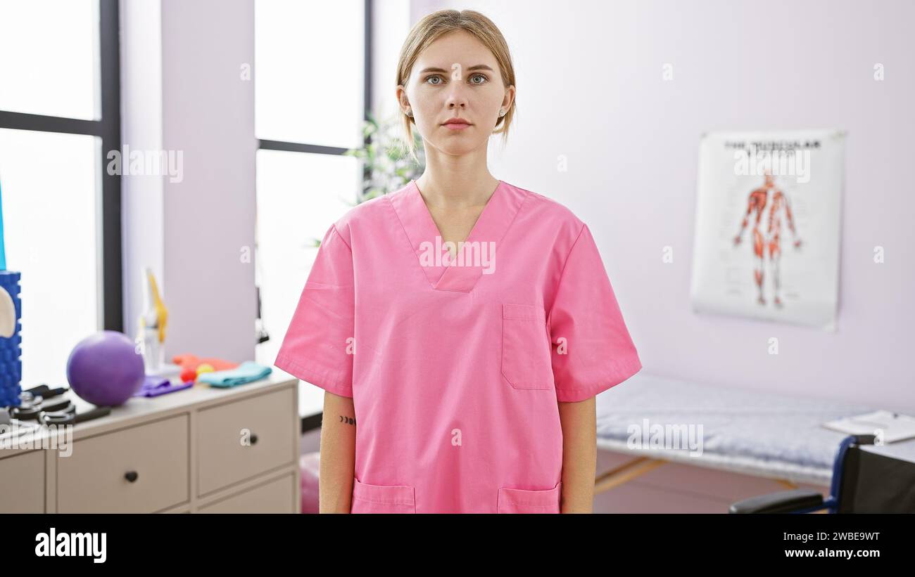 A caucasian woman in pink scrubs stands confidently inside a well-equipped rehabilitation clinic. Stock Photo
