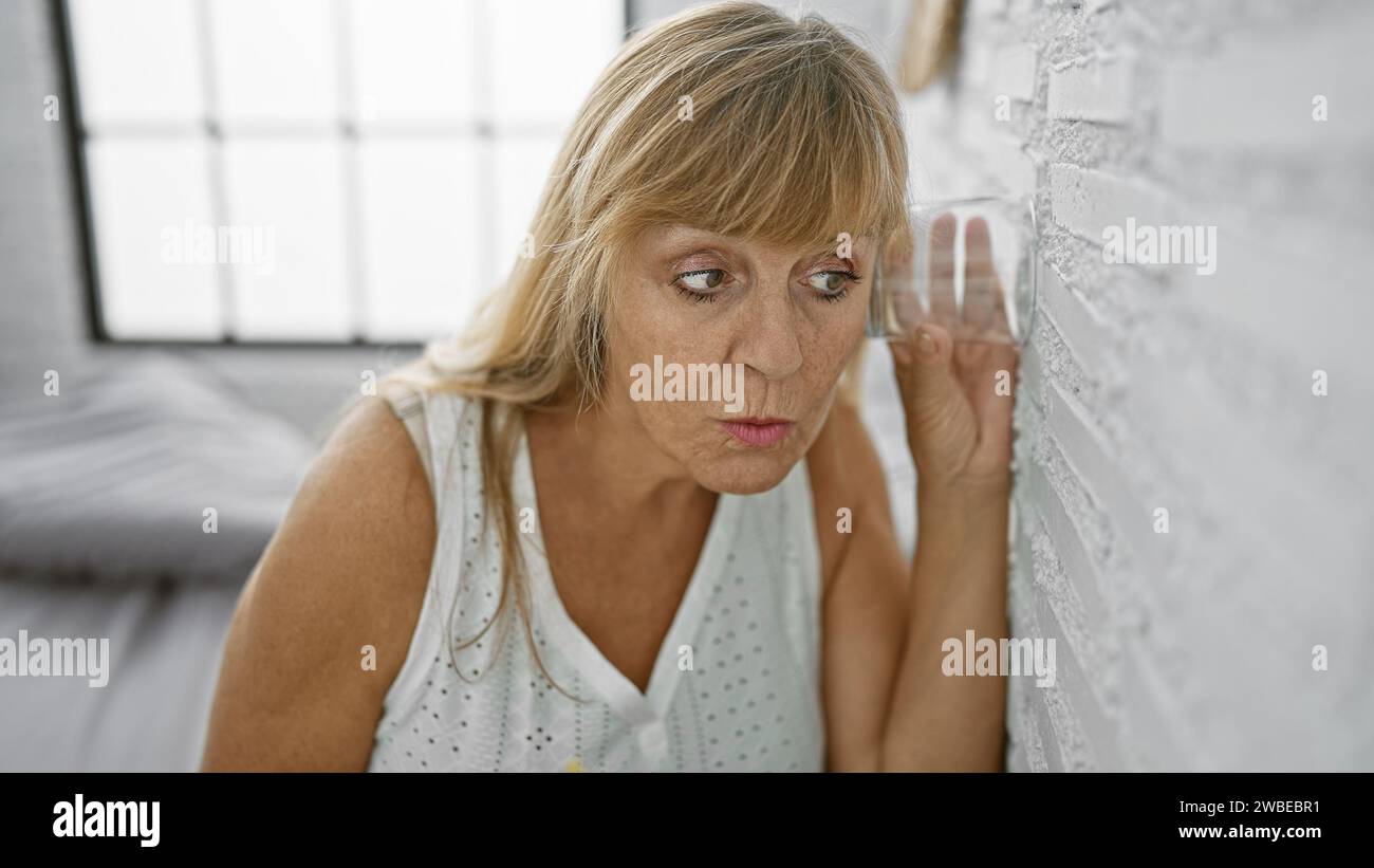 Curious middle age blonde woman eavesdropping, spying through bedroom wall with glass, indoor