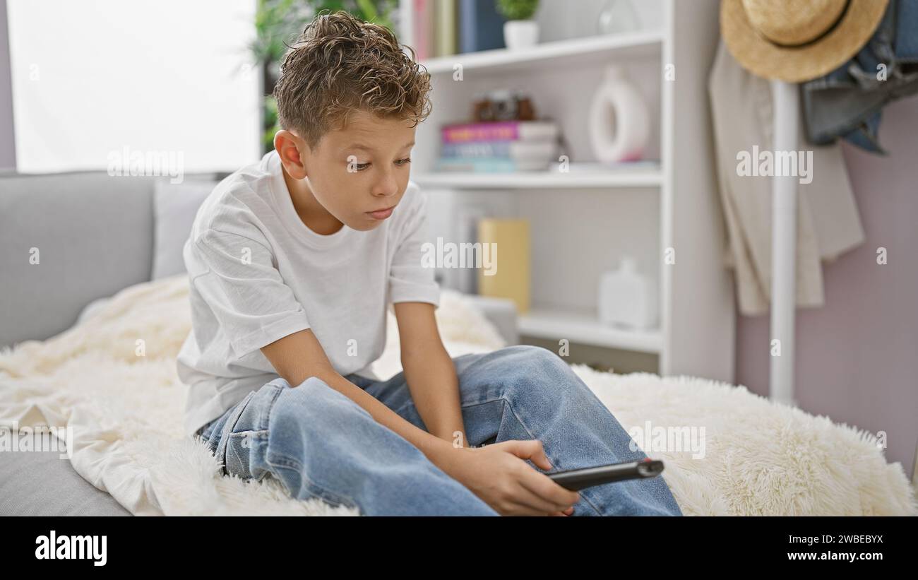 Adorable blond kid wearing a bored expression, comfortably hustling on the sofa at home, seeming serious while relaxing and watching tv in the living Stock Photo