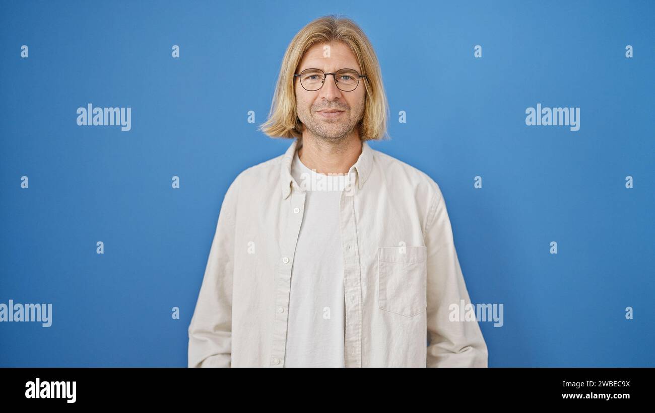 Handsome blond man with glasses and long hair wearing a casual shirt against a solid blue background looking at the camera. Stock Photo