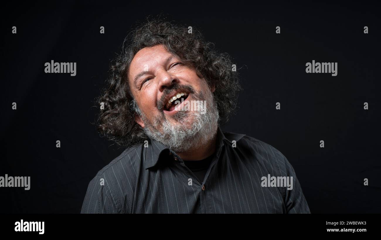 Portrait of latin man with white beard and black curly hair with funny expression, looking up, wearing black shirt against black background Stock Photo