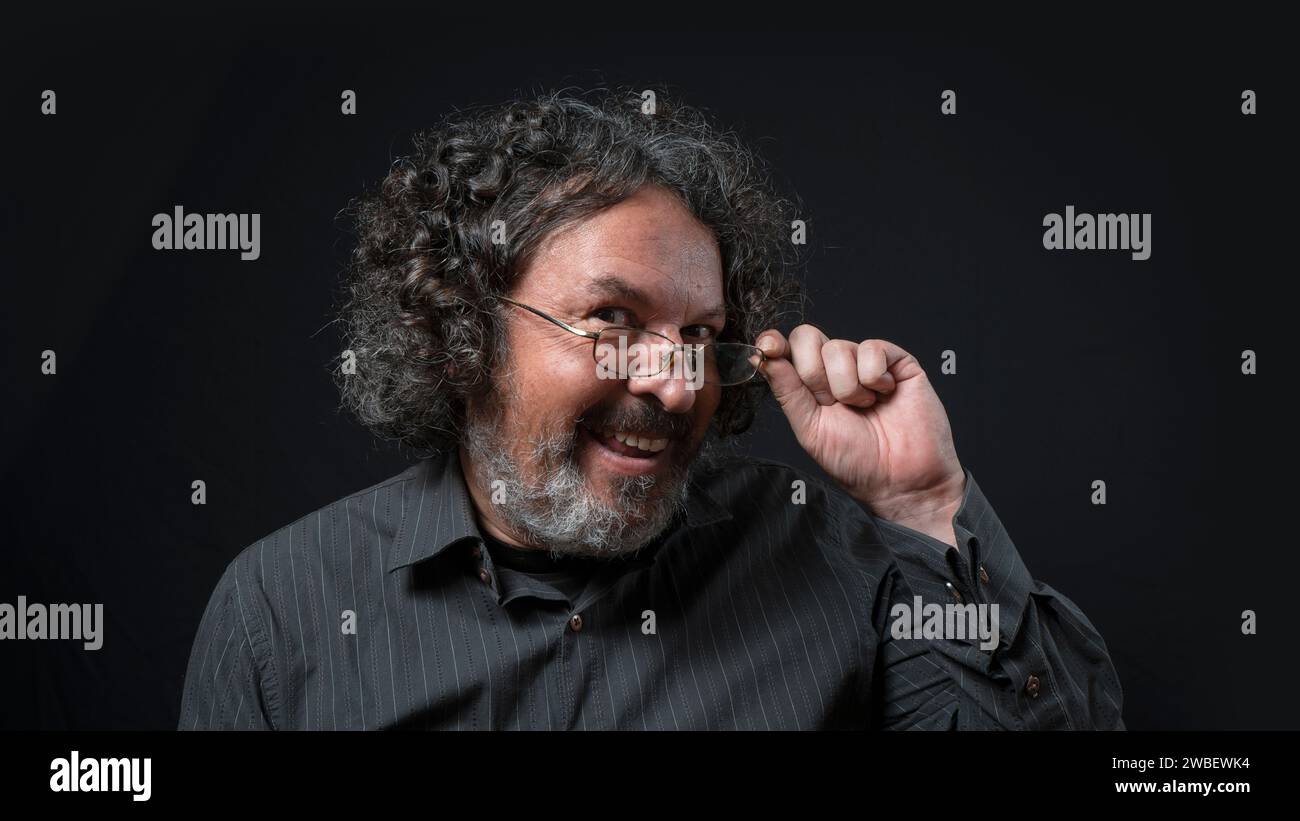 Portrait of latin man with white beard and black curly hair with flirtatious expression, holding his glasses, wearing black shirt against black backgr Stock Photo