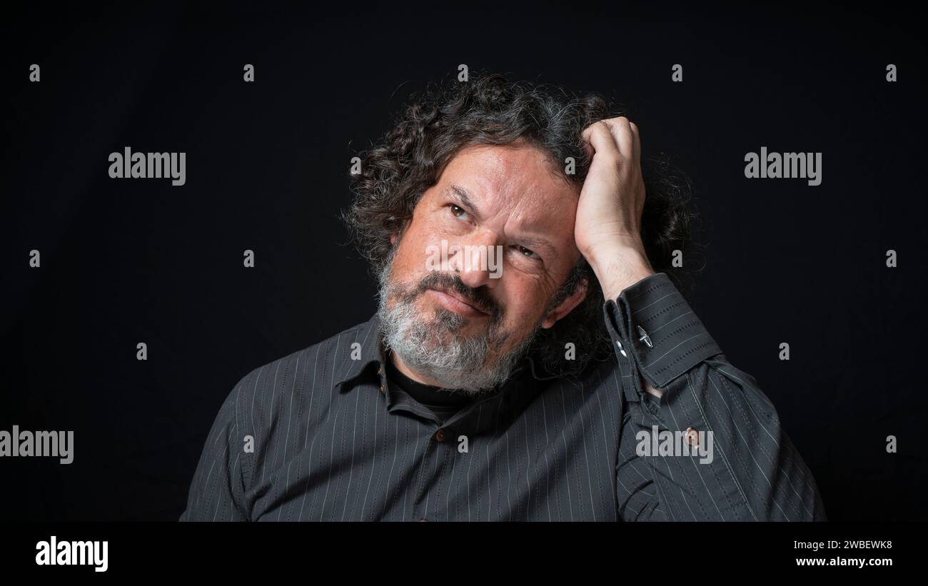Portrait of latin man with white beard and black curly hair with confused expression, scratching head with his hand, wearing black shirt against black Stock Photo