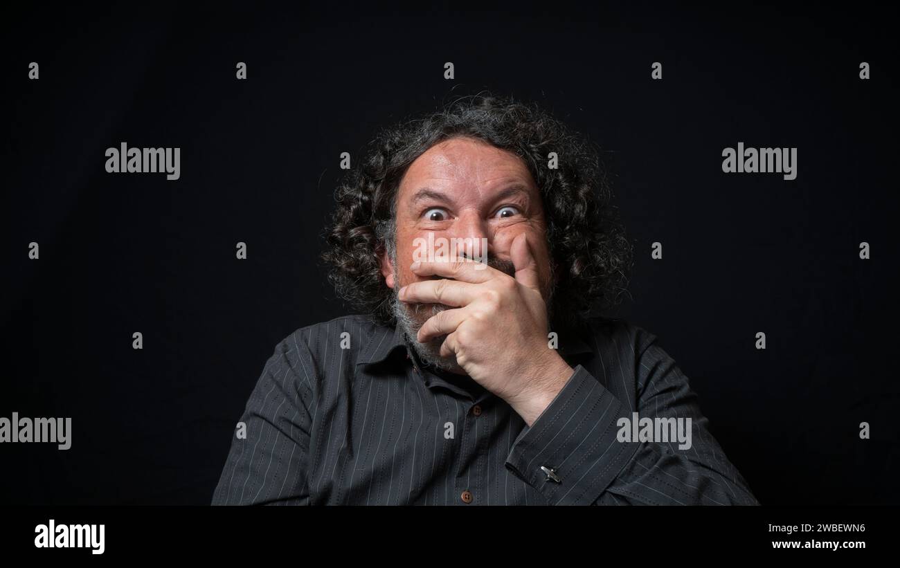 Portrait of latin man with white beard and black curly hair with funny expression, covering his mouth with hand, wearing black shirt against black bac Stock Photo