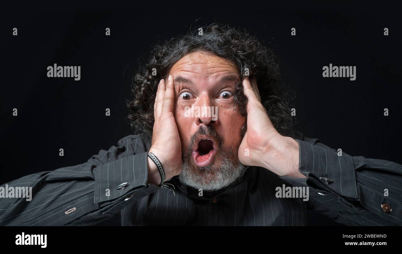 Portrait of latin man with white beard and black curly hair with surprised expression, hands on face, wearing black shirt against black background Stock Photo