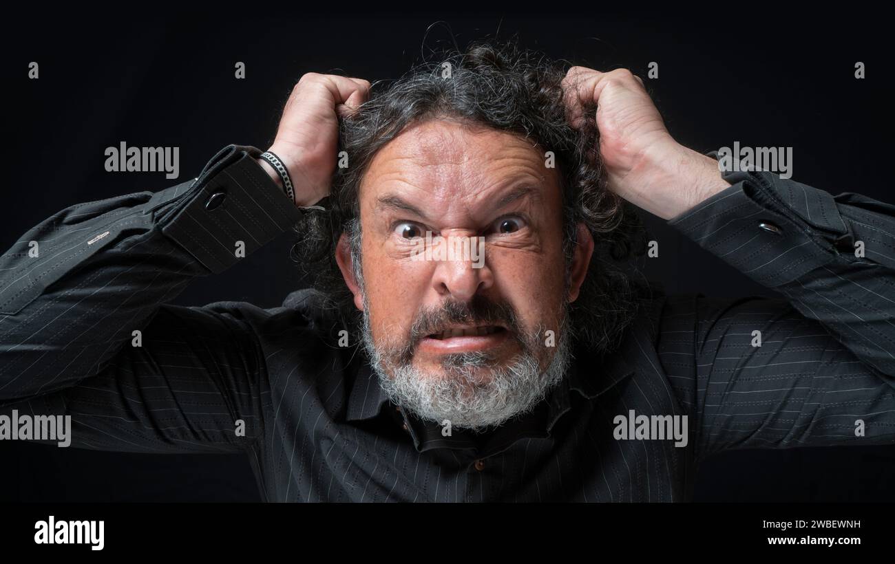 Portrait of latin man with white beard and black curly hair with very angry expression, grabbing his hair with hands, wearing black shirt against blac Stock Photo