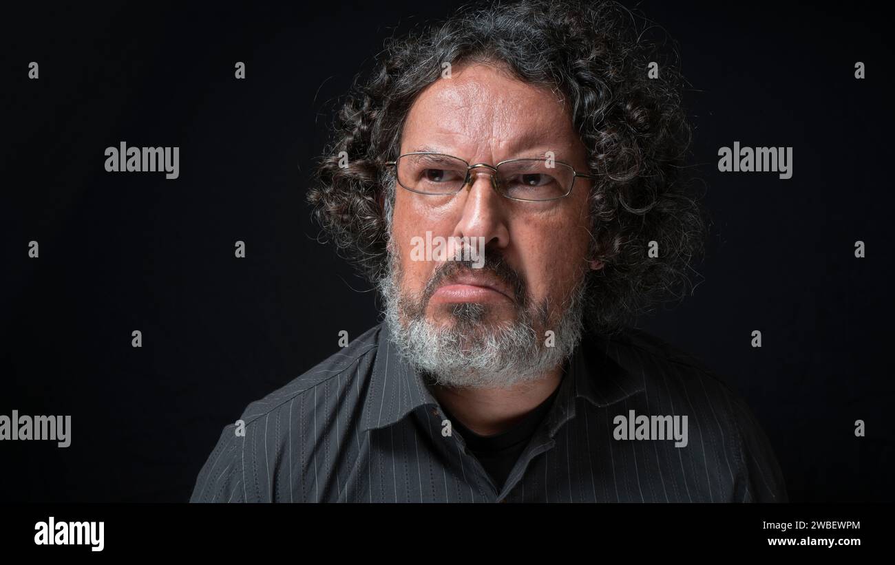Portrait of latin man with white beard and black curly hair with frustrated expression, grimacing with mouth, wearing black shirt against black backgr Stock Photo