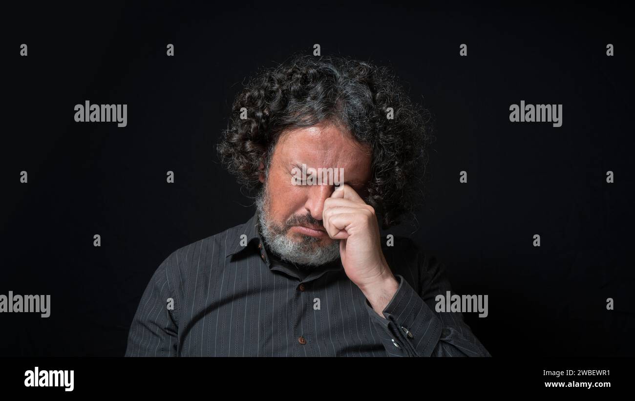 Portrait of latin man with white beard and black curly hair with sad expression, eyes closed and head down, wearing black shirt against black backgrou Stock Photo