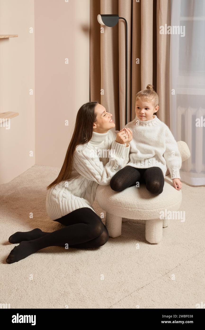 A young mother sits on the floor next to her little daughter sitting on a soft chair Stock Photo