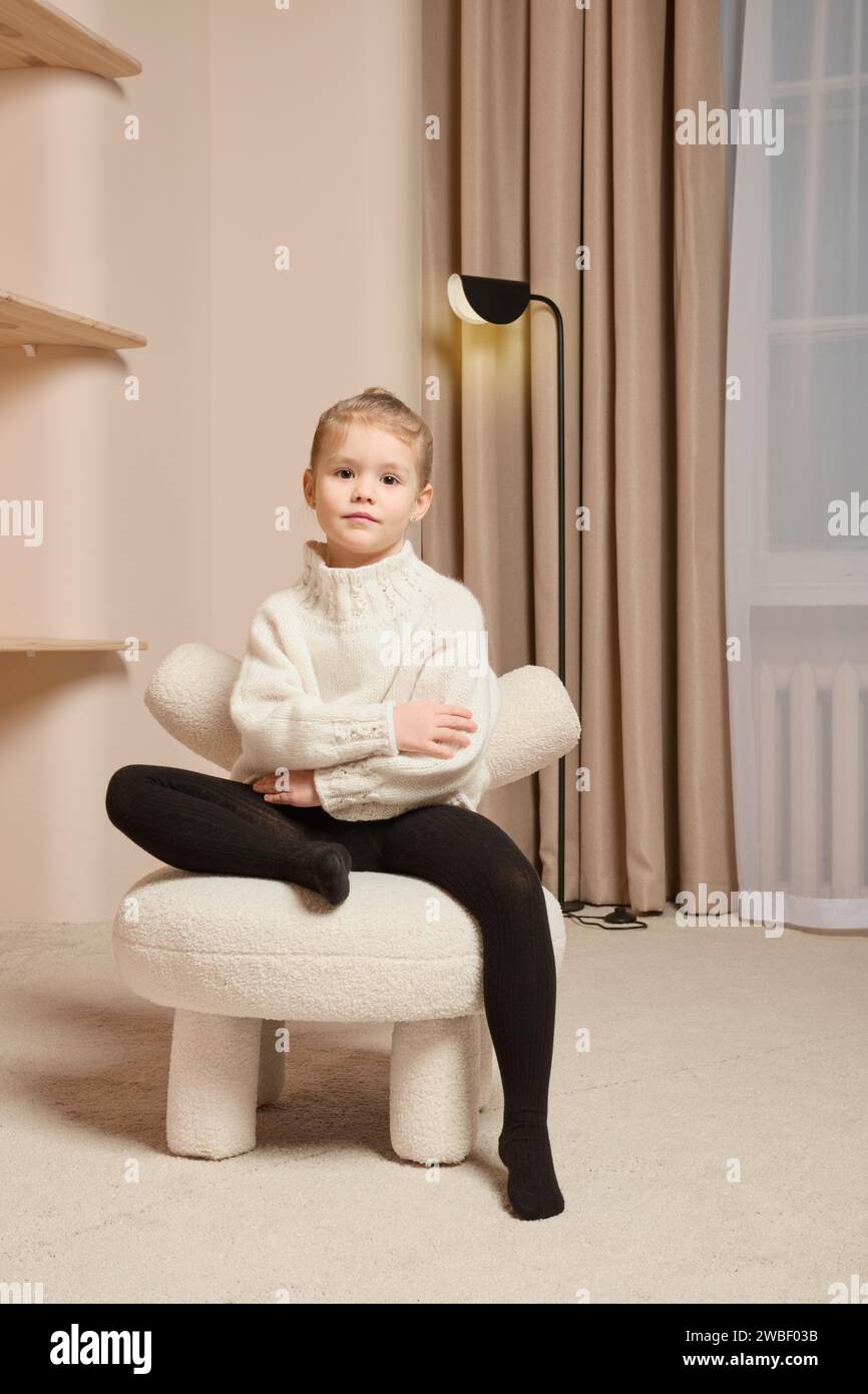 A little girl in warm sweater sits on a soft chair with one leg crossed and her arms crossed over her chest Stock Photo