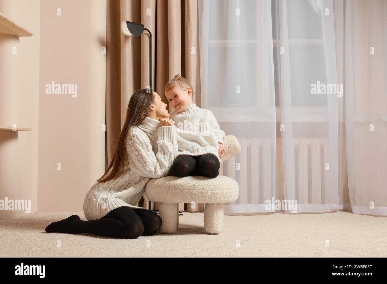 A young mother looks lovingly at her little daughter sitting on soft chair Stock Photo