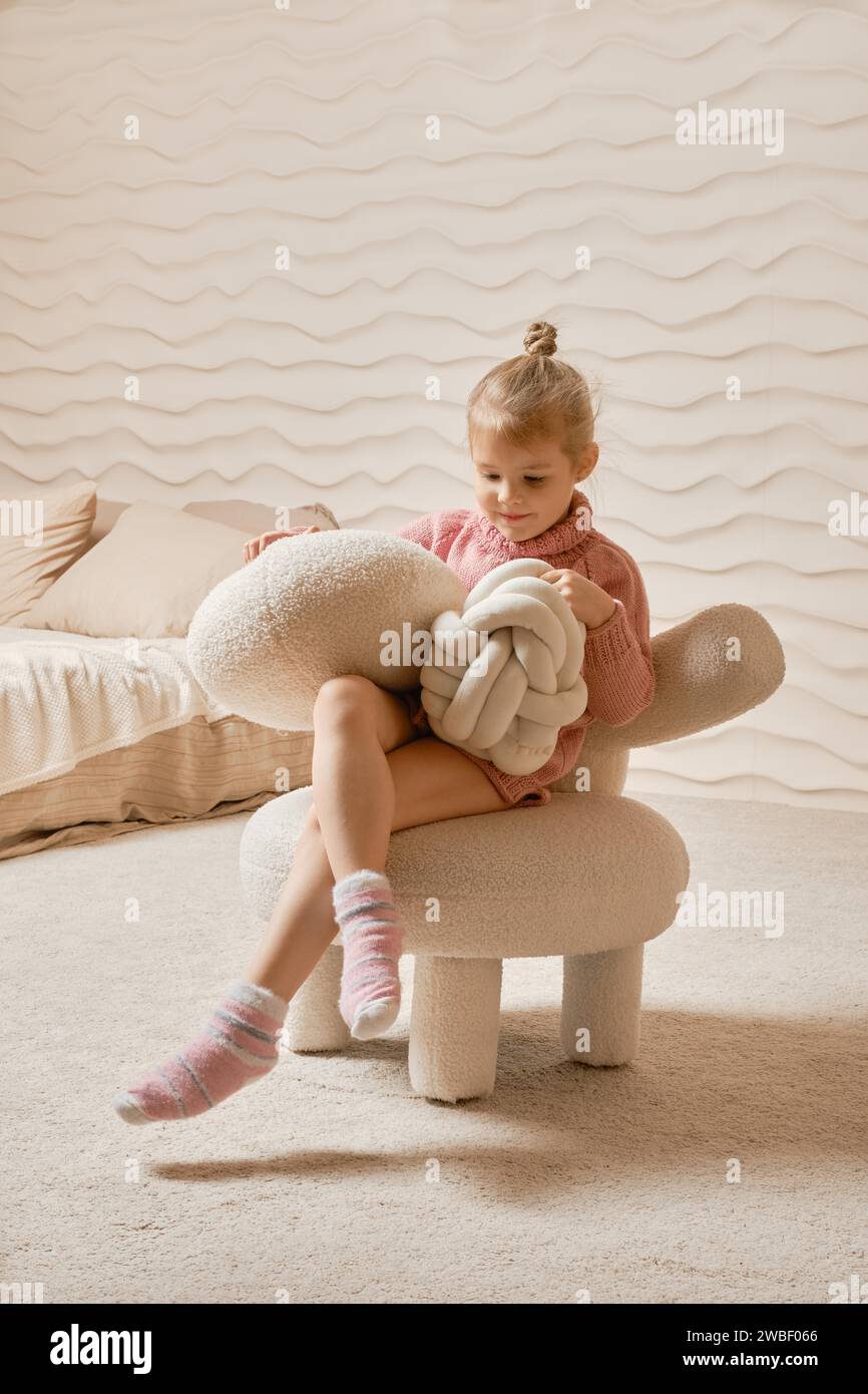 Little girl playing with stuffed pillows in her room, sitting on soft chair and knocking pillows Stock Photo