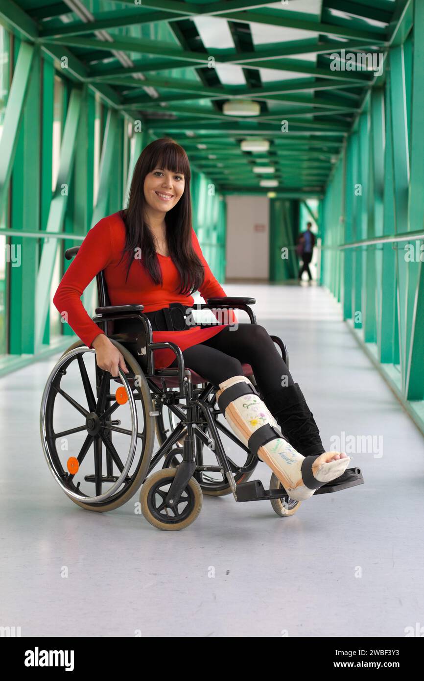 A young woman with a leg in plaster sits in a wheelchair and smiles, hospital Stock Photo
