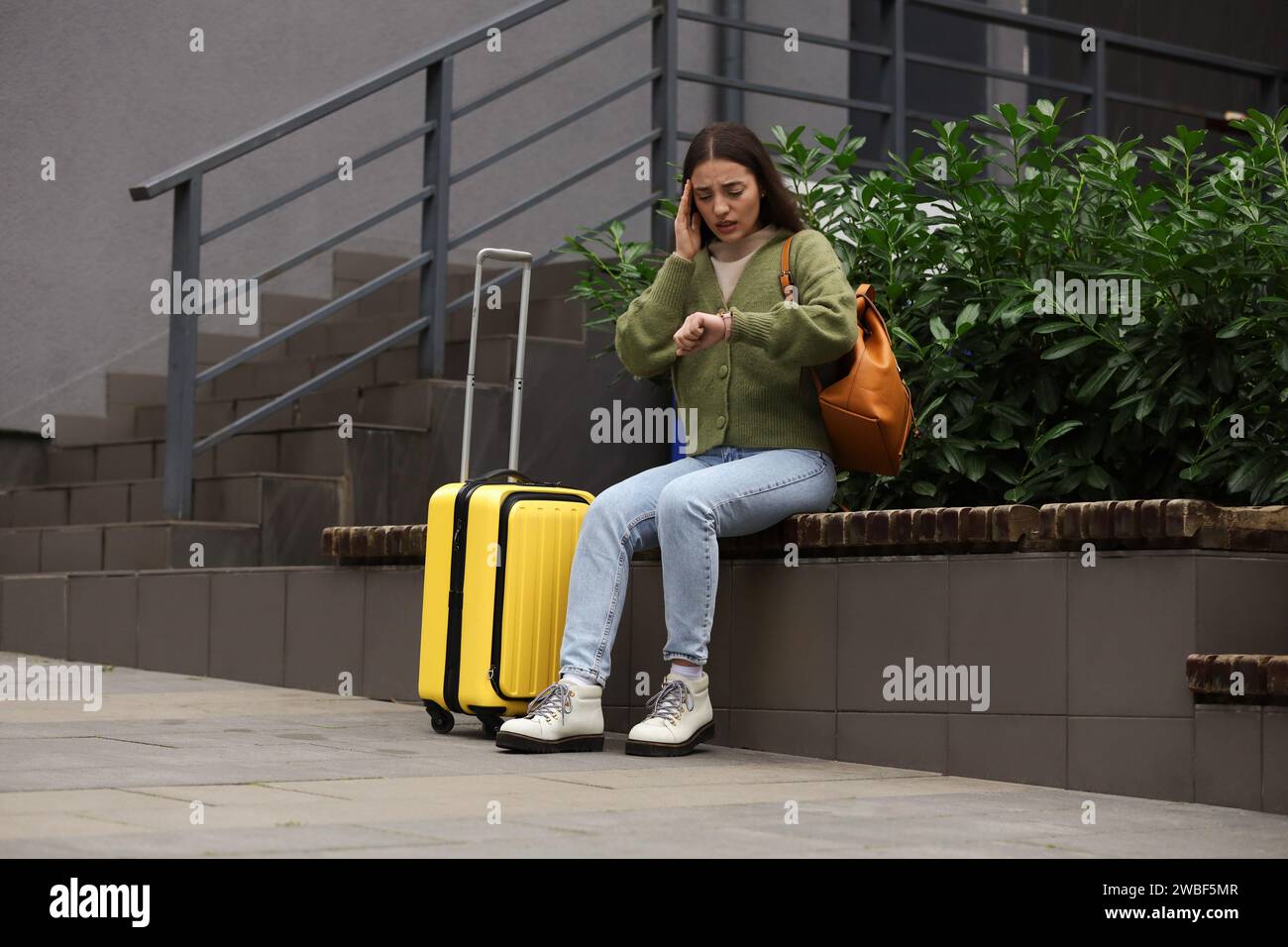 Being late. Worried woman with suitcase looking at watch on bench outdoors, space for text Stock Photo