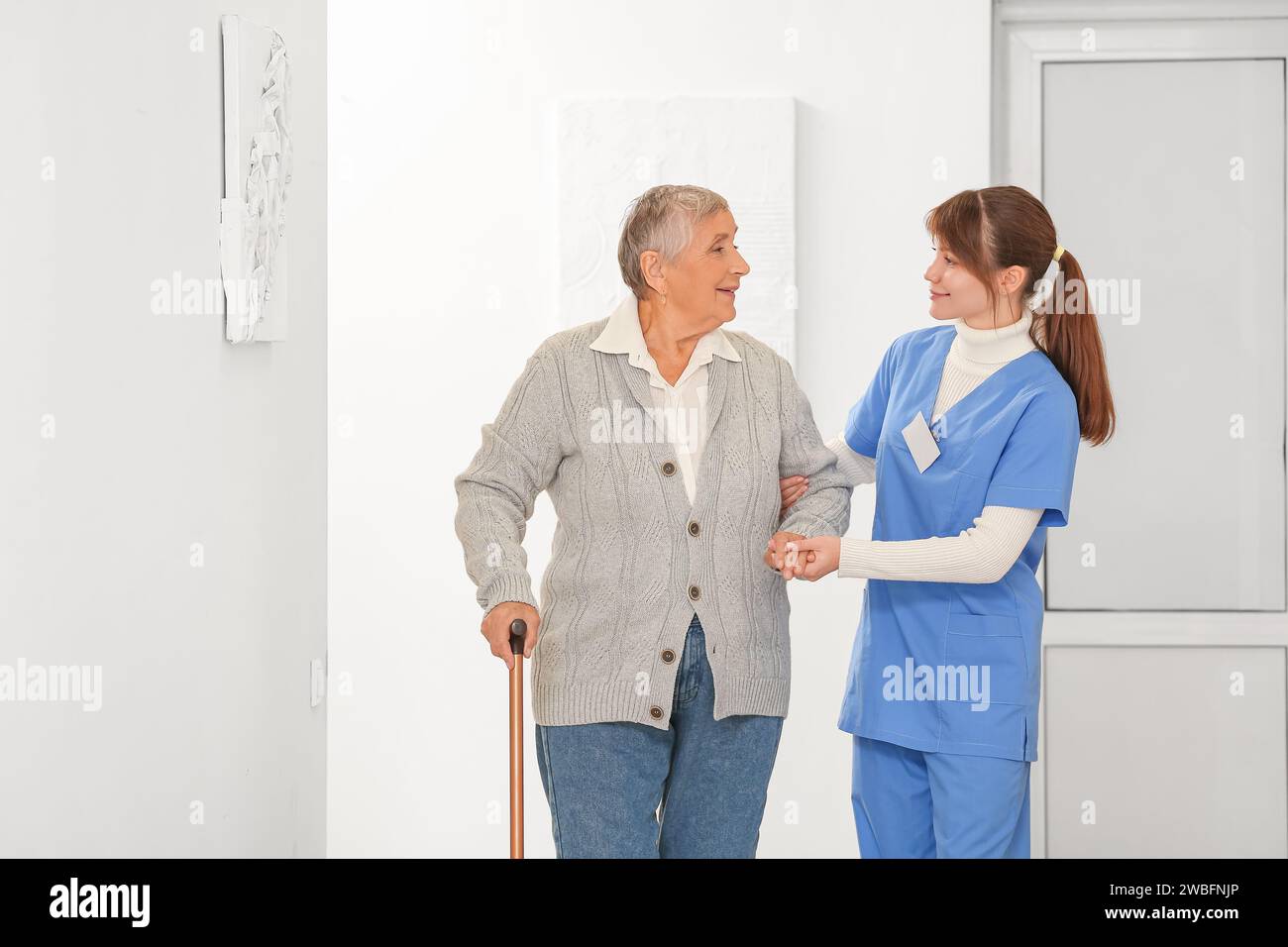 Senior woman with walking stick and caregiver in hallway Stock Photo