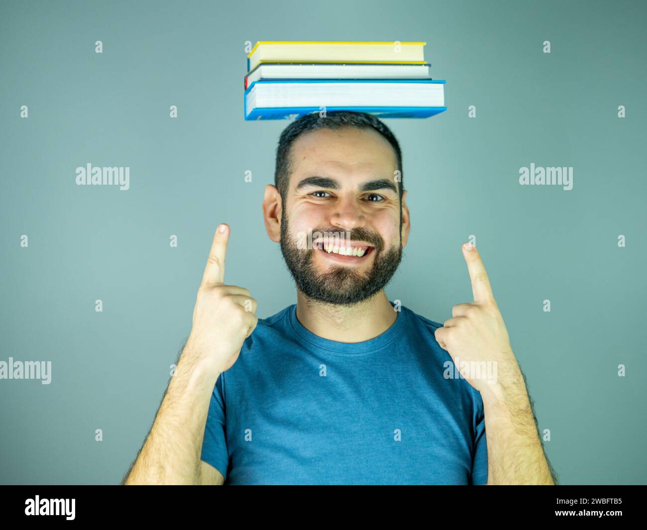 young bearded man with a pile of books on his head pointing up while looking at camera Stock Photo