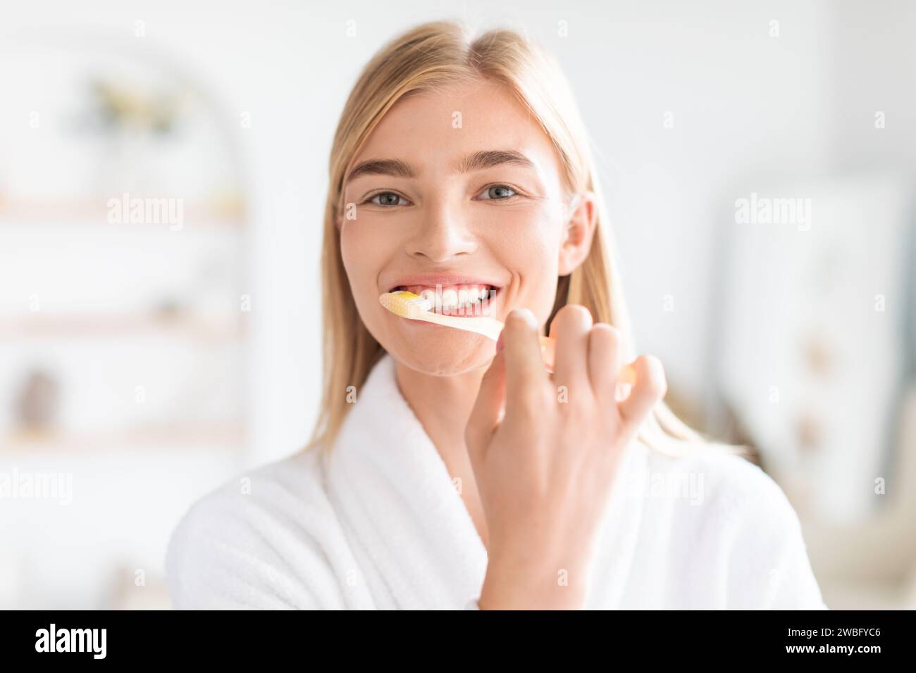 Portrait of pretty blonde woman attentively cleaning her teeth indoors ...