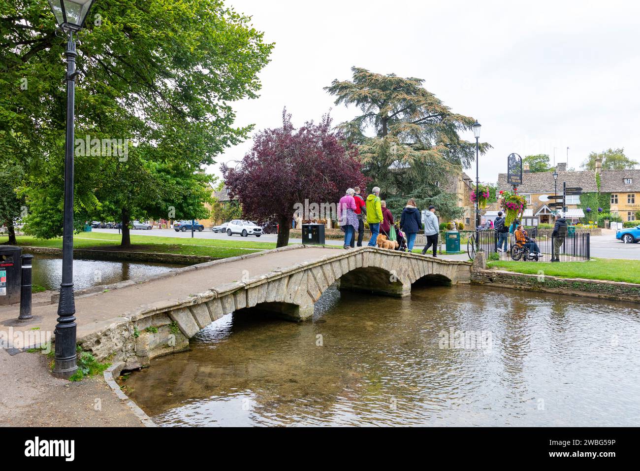 Cotswolds England , people with dogs walk across one of the low stone bridges in Bourton on the Water village, crossing the River Windrush, England,UK Stock Photo