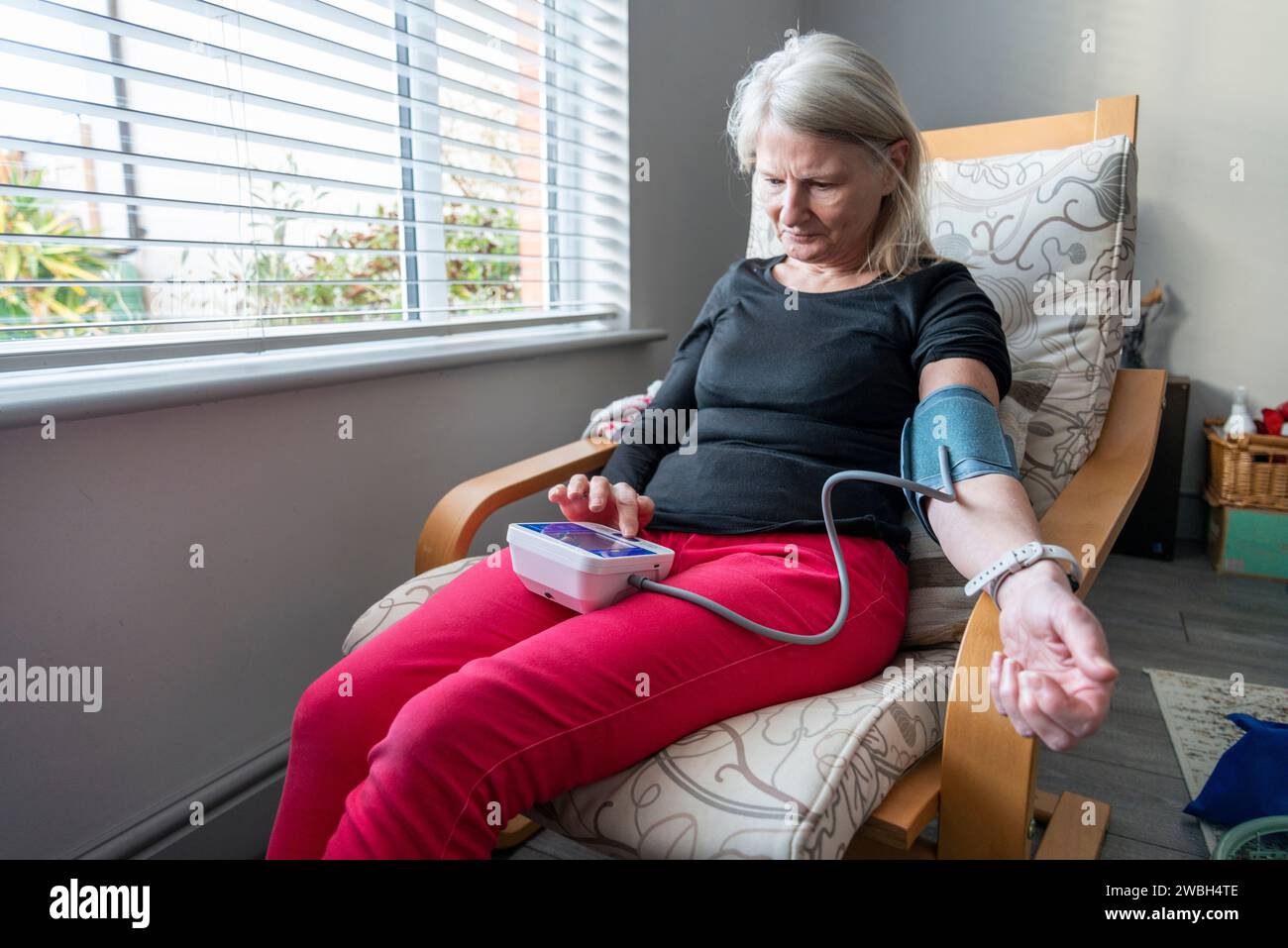 A health conscious female,sits in an armchair,in her house,using a blood pressure monitor to self-check her general physical condition,part of her hea Stock Photo