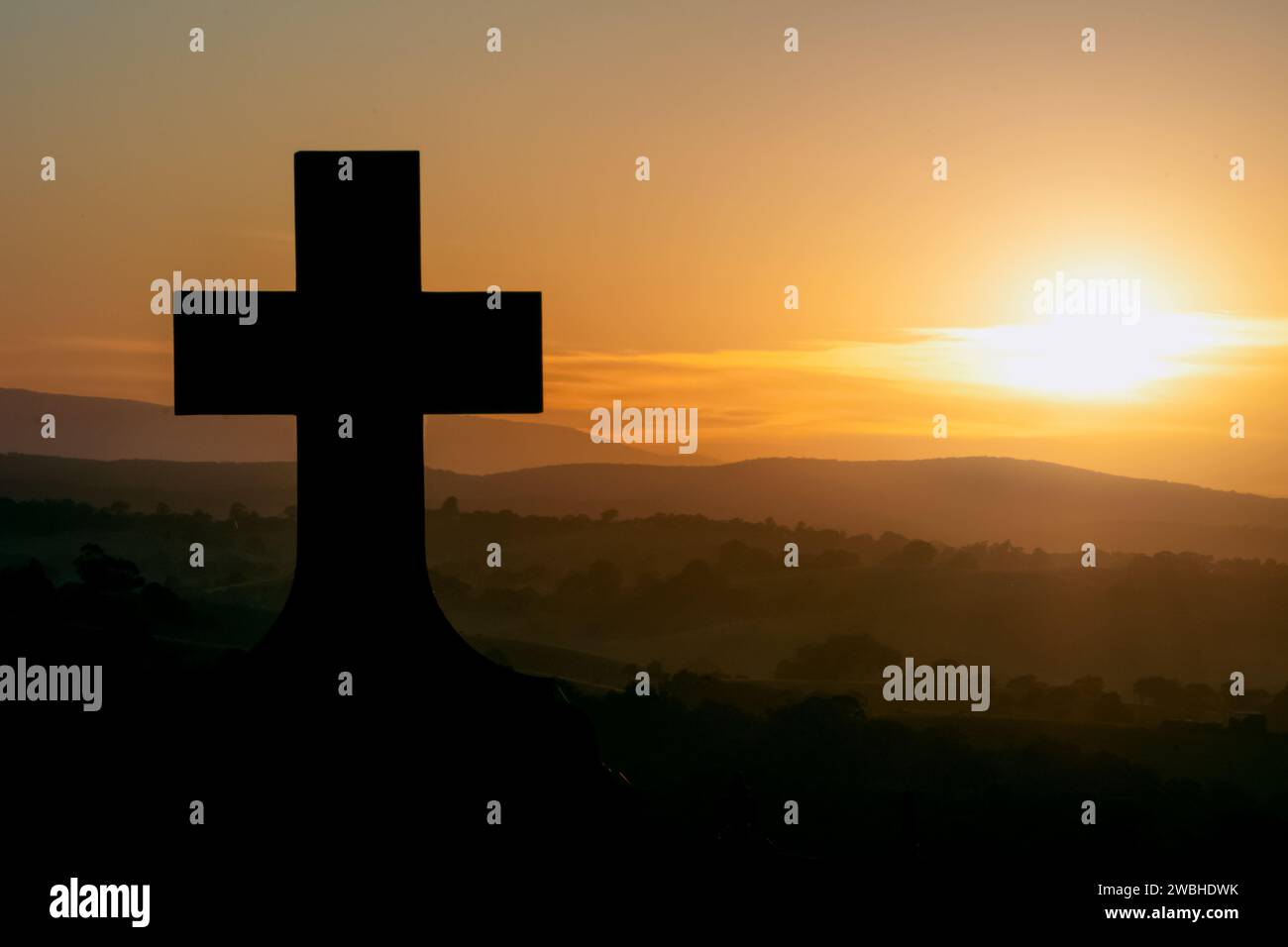 Silhouette of a Christian cross at sunrise, overlooking rolling hills Stock Photo