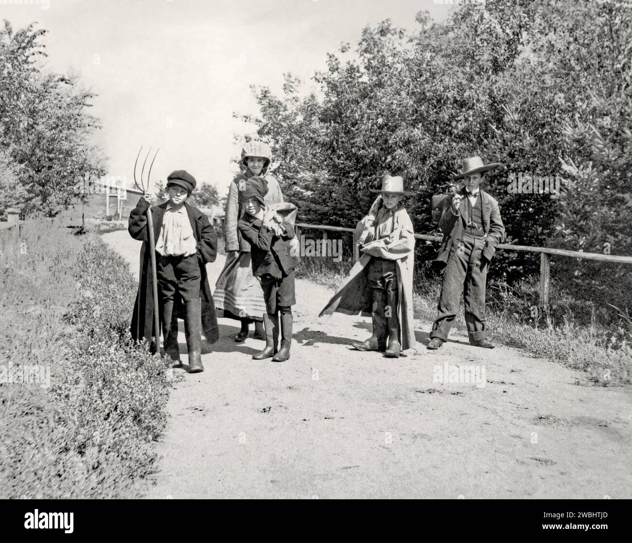 An old photograph of children out ‘tramping’ in the sunshine at Maple Grove Farm, Rosser, Manitoba, Canada in the early part of the 20th century. In the rural setting the four boys and a taller girl are having fun posing, dressed up wearing oversized, adult clothes and hats. The girl wears an upturned basket on her head. One boy carries a pitchfork. This is taken from an early photograph album – this photo was captioned as ‘Amateur Tramps’. Stock Photo