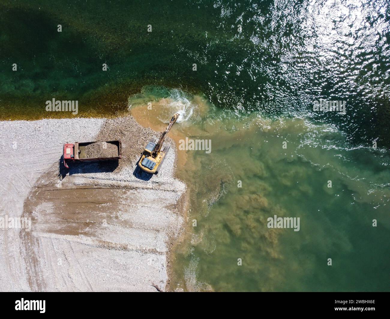 Excavator digging and loading sand into dump truck at river bank. Heavy machinery working at sand quarry. Aerial drone view of digger and truck. Stock Photo