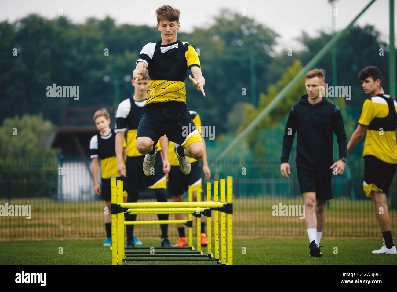 Football camp for youth. Teenage boys in training with a coach at grass pitch. Sports team preparing for the season. Young players jumping over hurdle Stock Photo