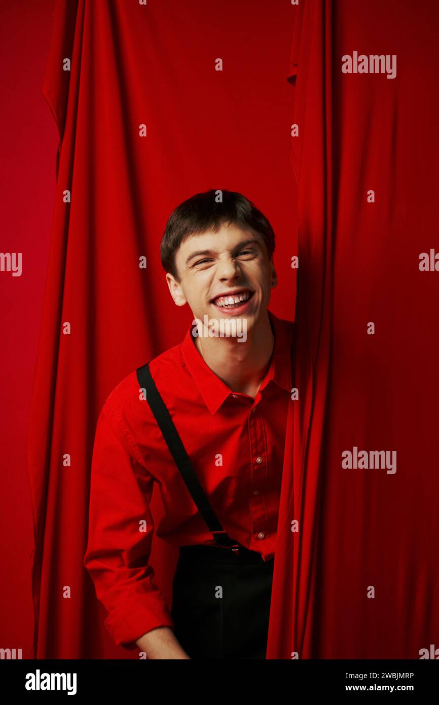 cheerful young man in suspenders smiling while hiding behind red vibrant curtain, merriment Stock Photo