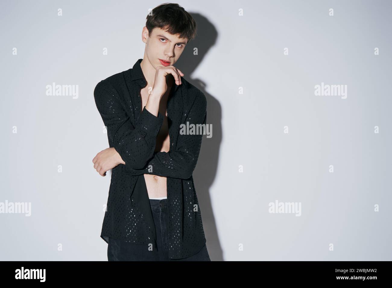 handsome young man in shiny black open shirt with hand near face posing on grey background Stock Photo