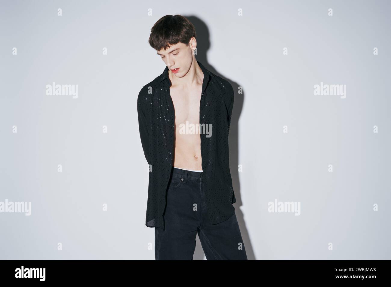 confident young man in shiny black open shirt posing with hands behind back on grey background Stock Photo