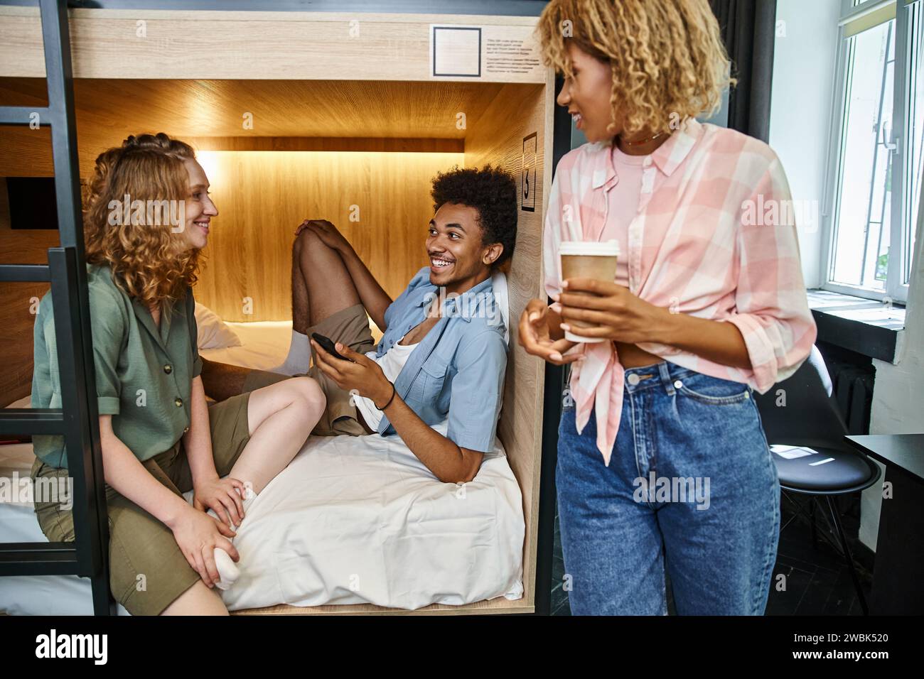 joyful african student with smartphone on double-decker bed near female friends in youth hostel Stock Photo