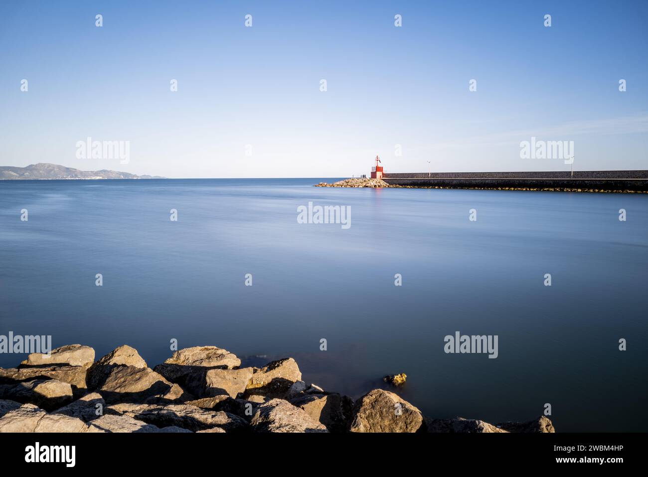 Far away red lighthouse, surrounded by rocks and a calm blue sea. Stock Photo