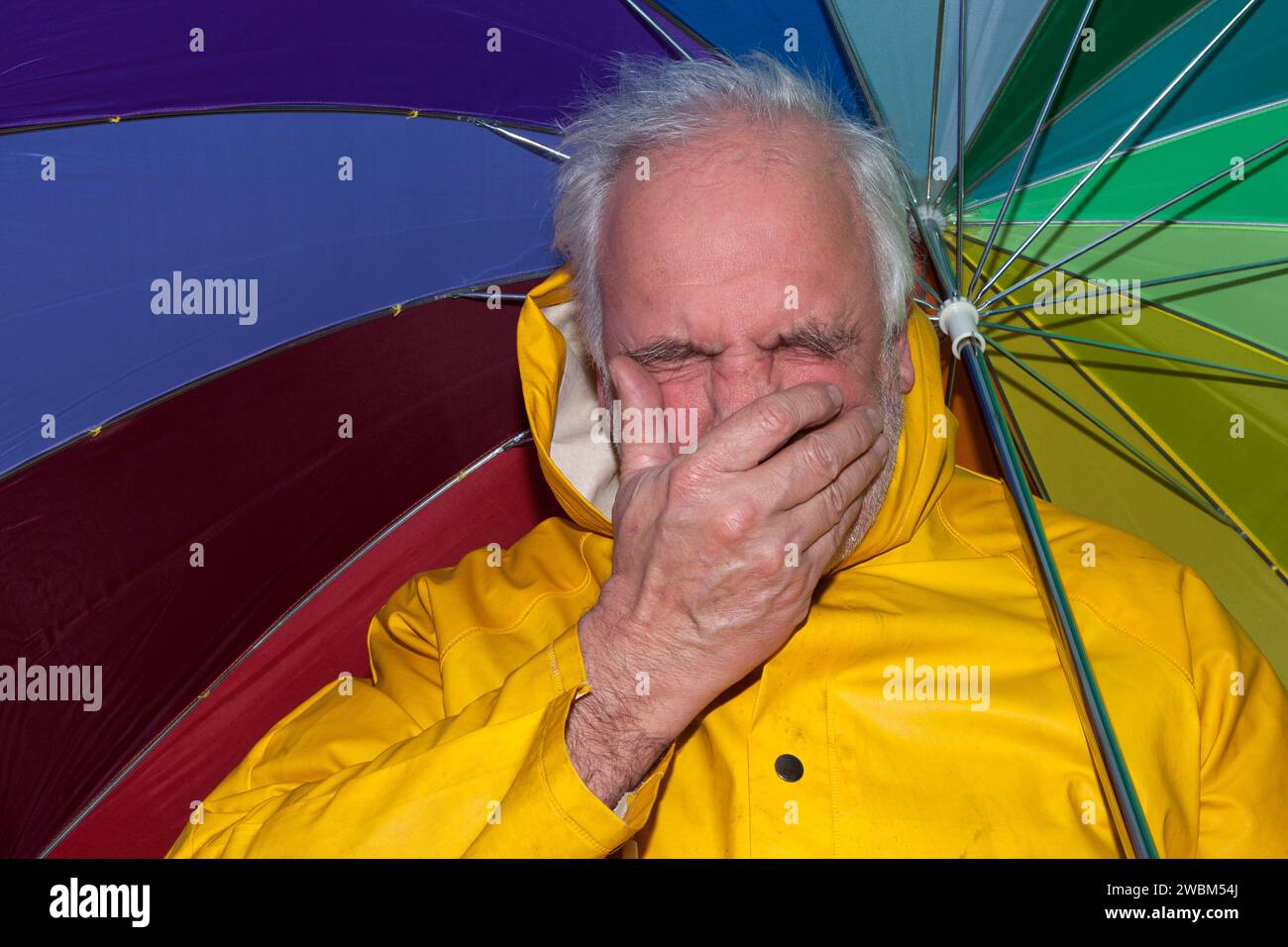 Man in yellow rain jacket and umbrella sneezing in rainy weather, showing cold symptoms. Realistic representation of rainy days and health. Stock Photo
