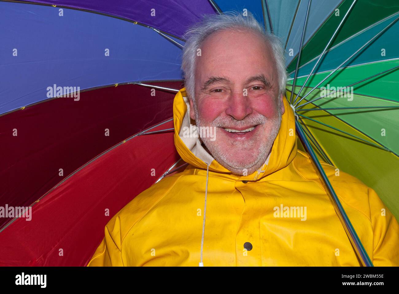 In a good mood despite bad weather, a smiling senior in a yellow rain jacket and a colorful umbrella radiates joy. Stock Photo