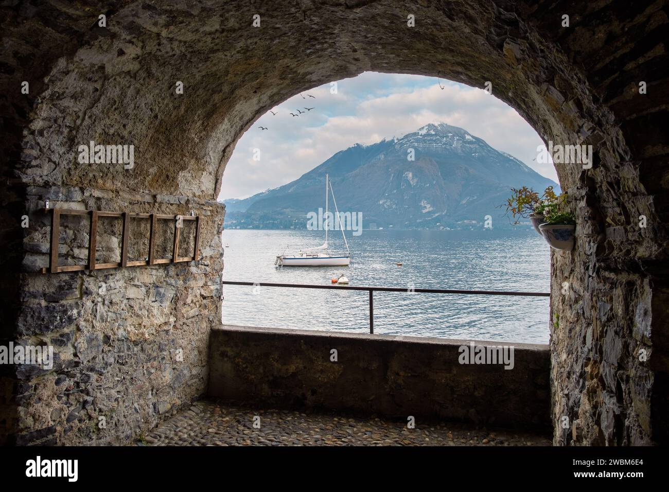 Sailboat docked in Varenna at winter time framed by a stone arch shaped window. Mountains in the distance, Lake Como, Varenna, Lakes Region, Lombardy, Stock Photo