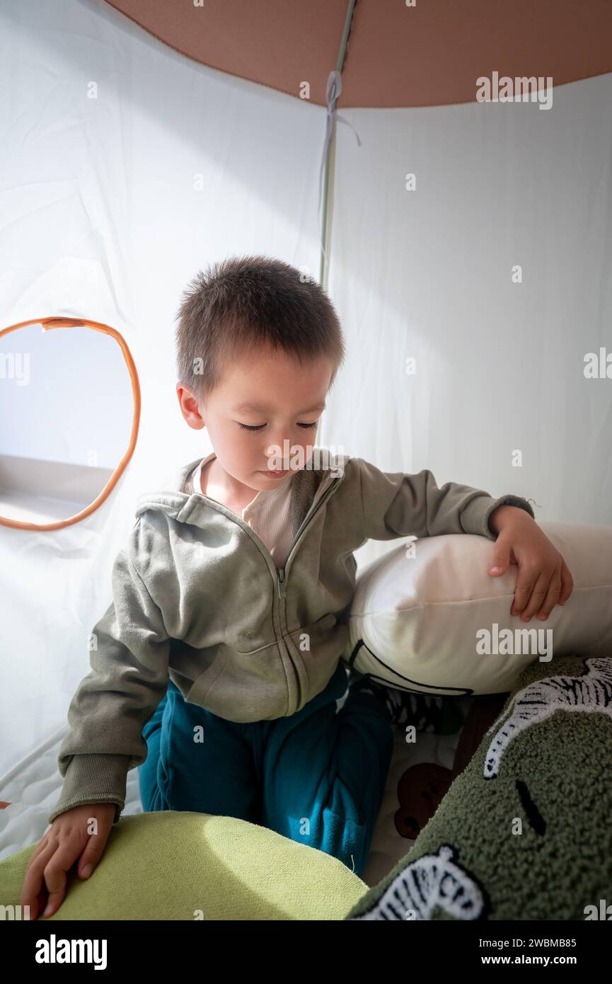 Adorable two and a half year old boy playing among the soft pillows in the kids tent. Boy is having fun while making himself a comfortable bed of pill Stock Photo