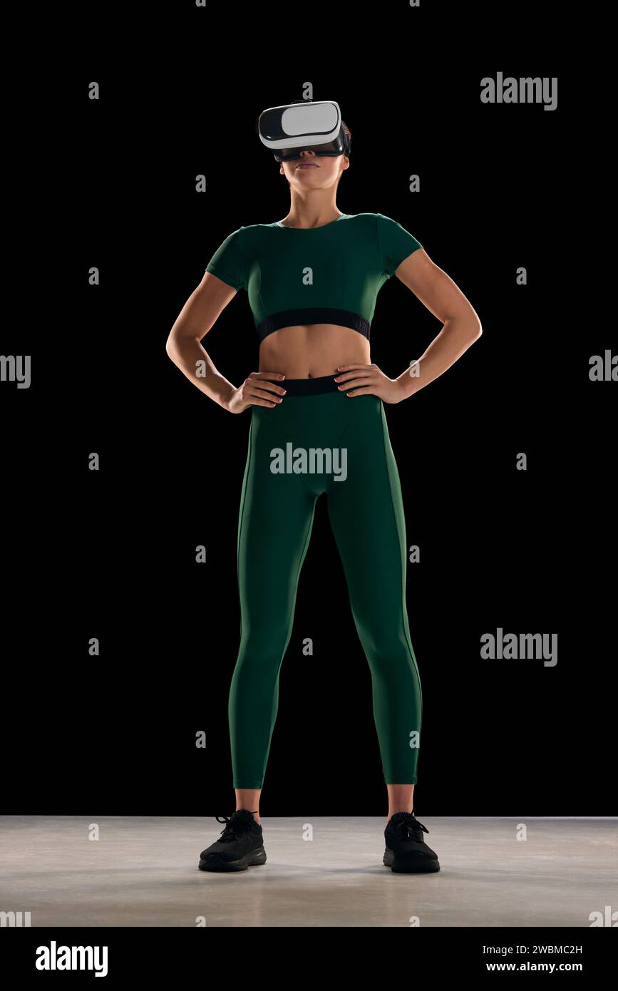 Full-length image of young woman with slim body in sportswear wearing VR glasses against black studio background. Online sport program Stock Photo